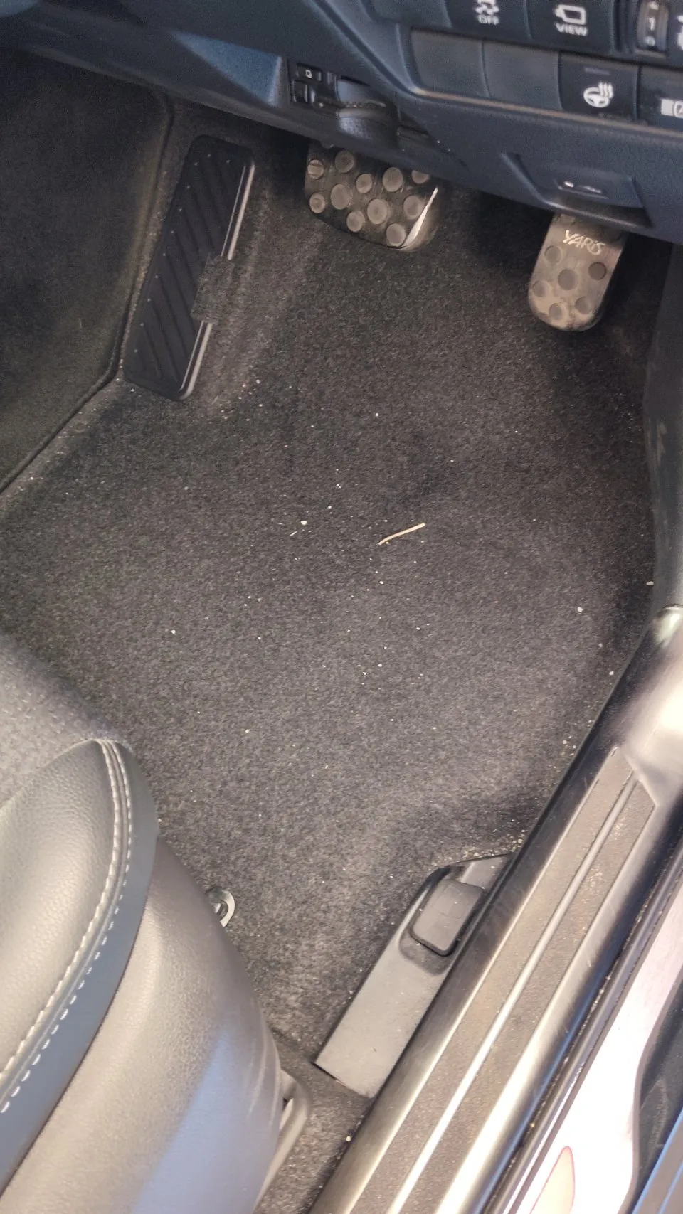トヨタ ヤリスクロスの施工前。運転席の足元を写した写真で、ペダルとその手前のカーペットに白っぽい細かい粒が点々と散っている
