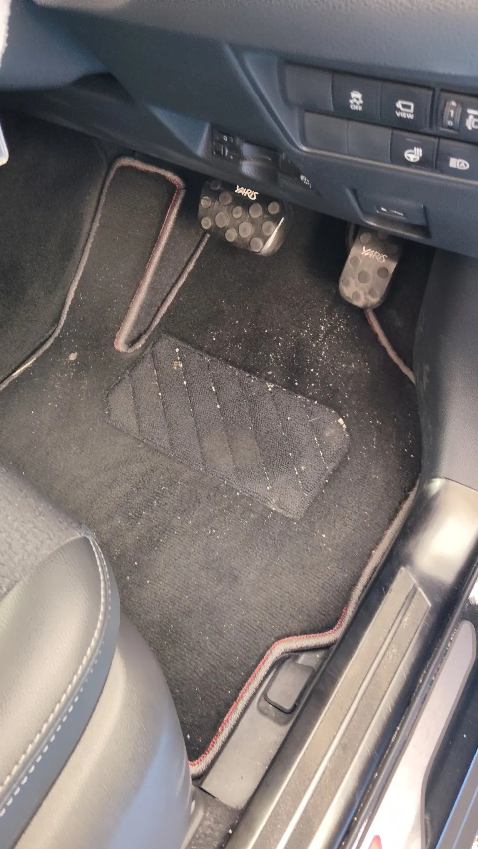 トヨタ ヤリスクロスの施工前。運転席の足元のフロアマットを写した写真で、ふちに赤いステッチが入ったマットの上に白っぽい細かい粒が散っている