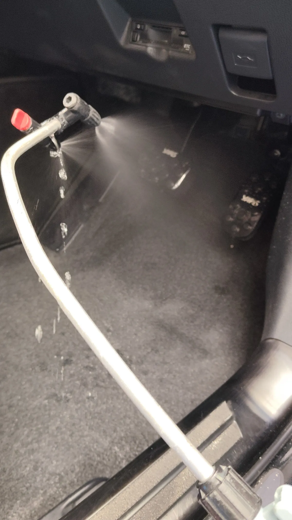 トヨタ ヤリスクロスの施工中。運転席の足元に向けて、赤いハンドルの付いた細い金属のノズルから霧状の洗浄液を噴射している
