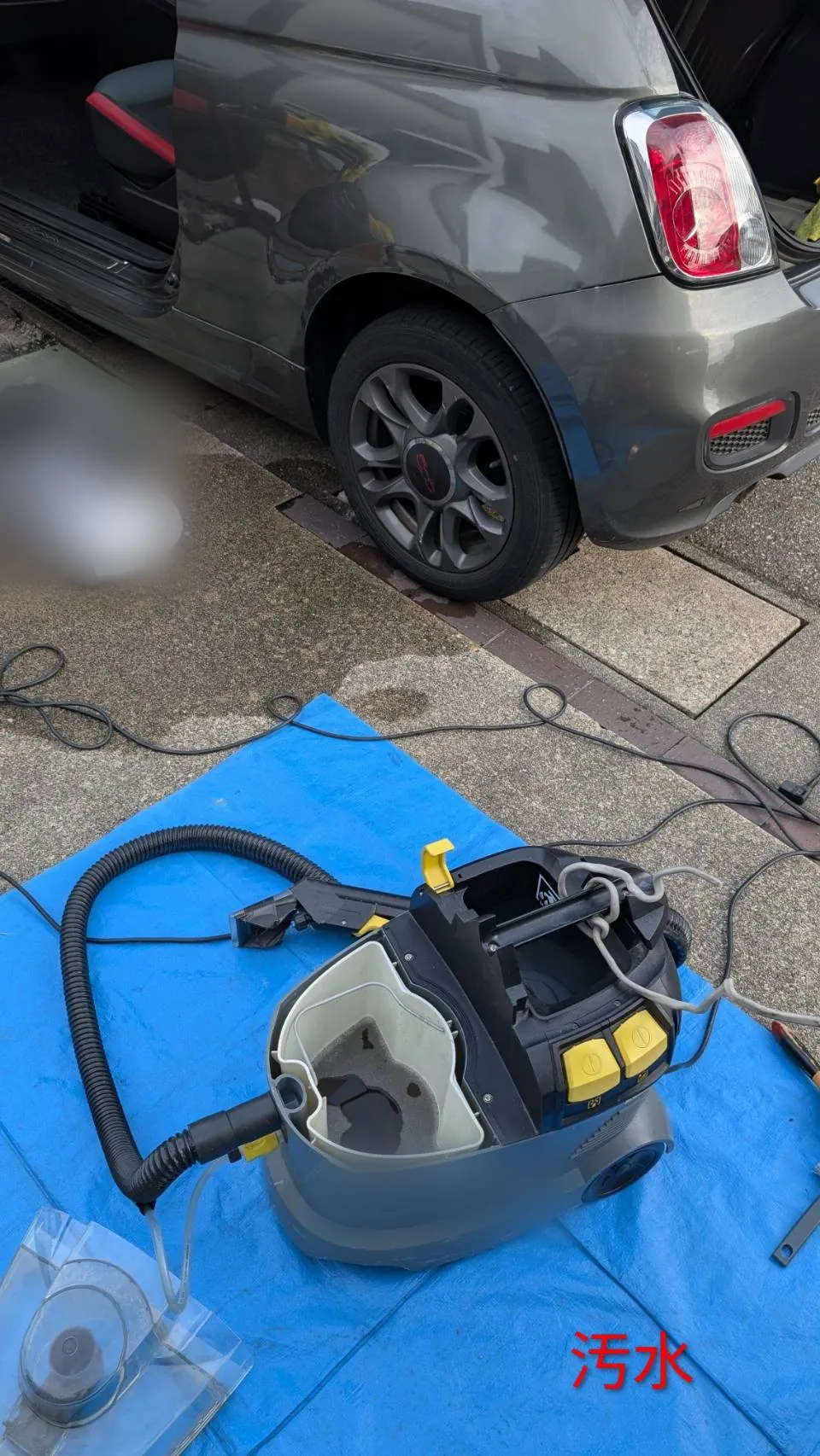 屋外の青いシートの上に置かれた機材のタンクを開けたところ。中に濁った水が残っている。奥に車両の後部が見える