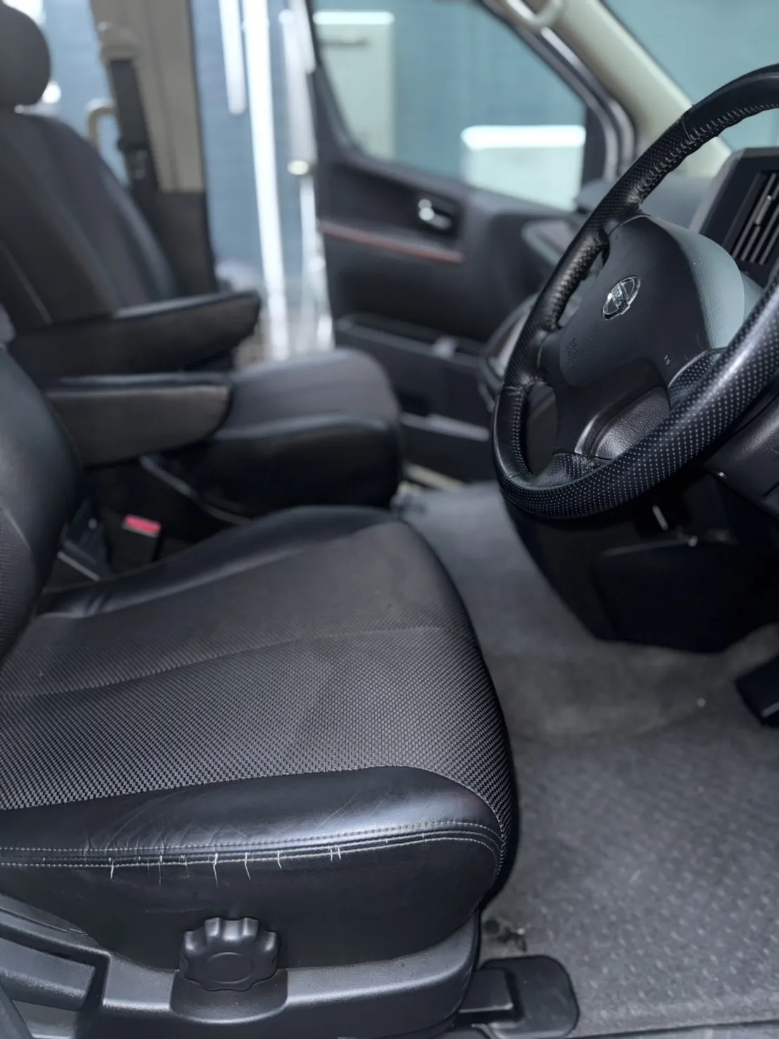 日産エルグランドの運転席まわり。座面の中央は細かい織り目のある生地で、周囲は黒い革調の素材。ステアリングは黒い革巻きで、中央に日産のエンブレム。足元はグレーのフロア。作業前の車内。