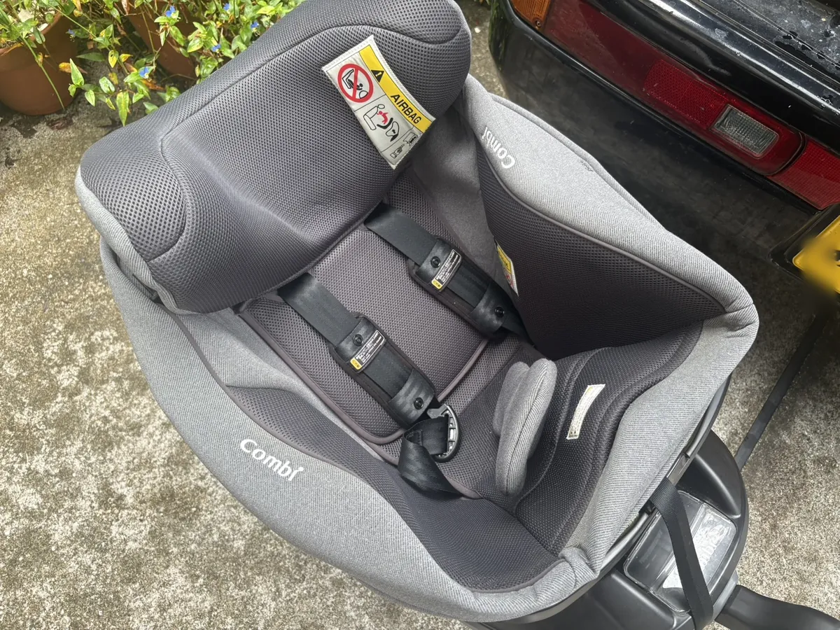 車から外したCombiのチャイルドシートをコンクリートの地面に置いたところ。グレーの生地で、肩2本と股1本の5点式ハーネスが付いている。背もたれの上部にAIRBAGの警告ラベル。左に植木鉢と植物、右に黒い車のテールランプ