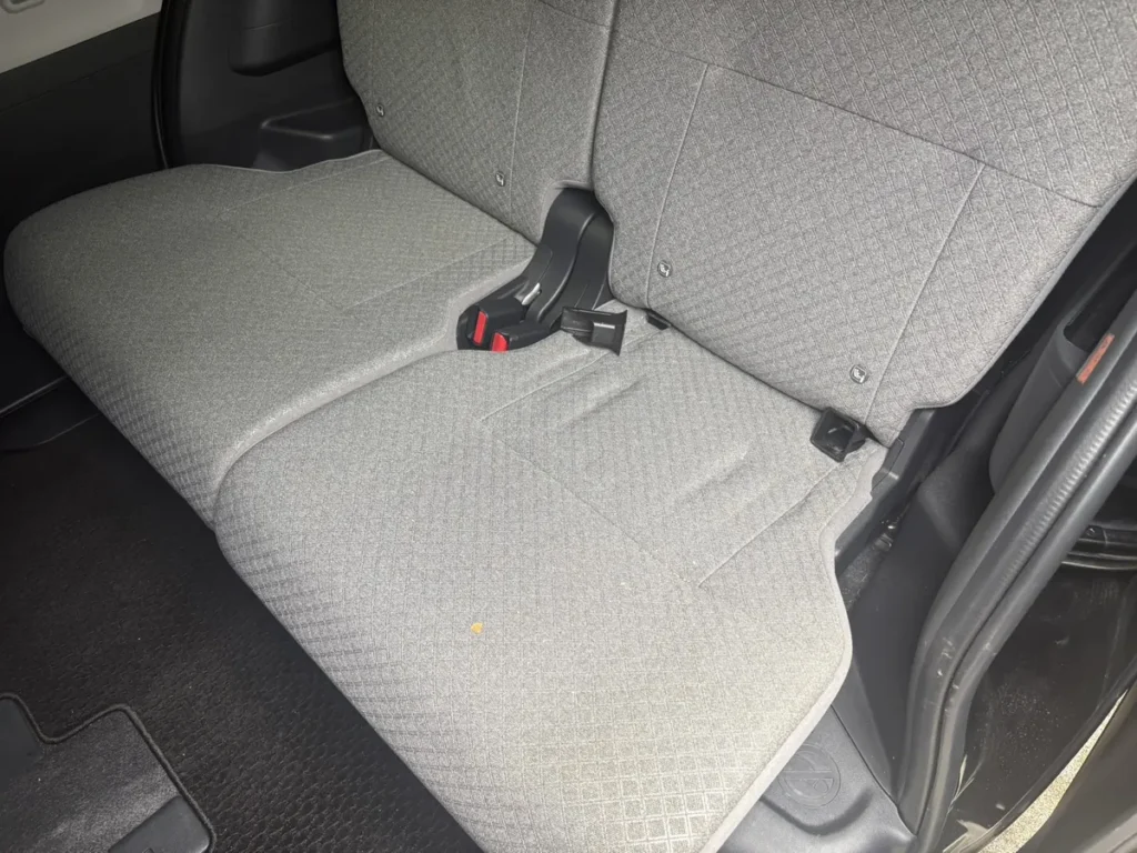 タントの後部座席を開けたドアの外から見たところ。グレーの布のシートで細かいダイヤ型の柄が織り込まれている。中央にシートベルトのバックル、座面の手前寄りに黄土色の小さな点がひとつある。床は黒いマット