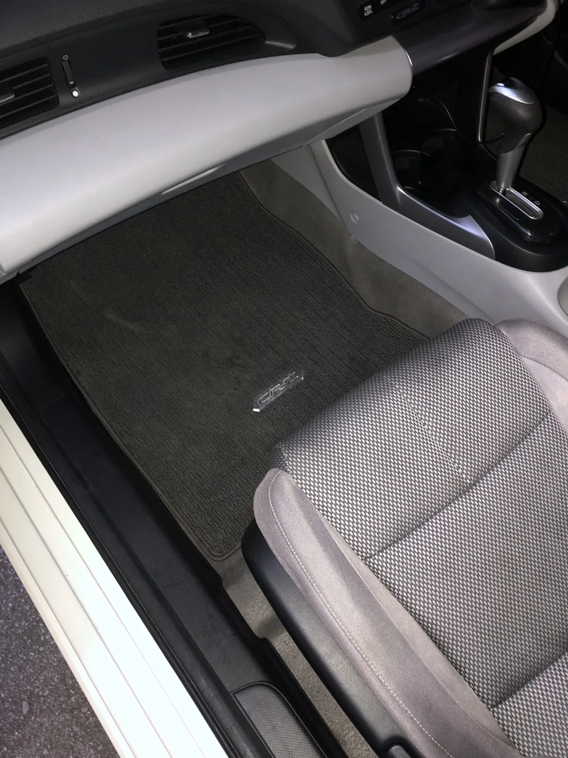 作業後のホンダ CR-Zの助手席の足元。助手席のドアを開けて外から撮ったところ。黒いフロアマットにCR-Zの銀色のロゴプレートが付き、マットの毛足がそろっている。左に白いサイドシル、右上にシフトレバーとオートマのセレクター、右下に助手席の座面が写っている