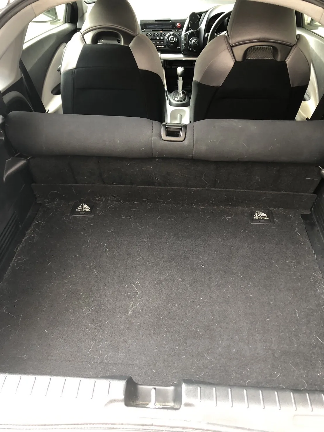 作業前のホンダ CR-Zの荷室。後ろのハッチを開けて外から撮ったところ。床も後席の背もたれの裏も黒い起毛のカーペット地で、床の全面に白い毛が散らばっている。床の奥に、TOP TETHERと書かれた黒い樹脂のカバーが左右に1つずつ付いている