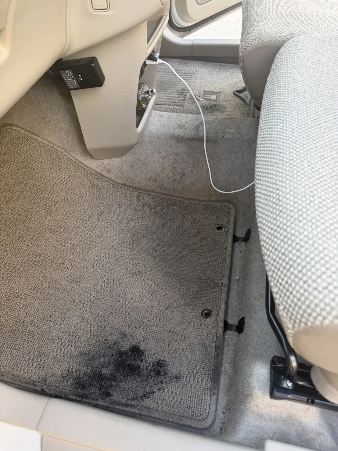 助手席の足元。グレーの細かいループのフロアマットの、ドア側の縁の近くに黒い跡が広がっている。マットの奥の床にも薄い黒ずみが点々と出ている。内装はベージュ
