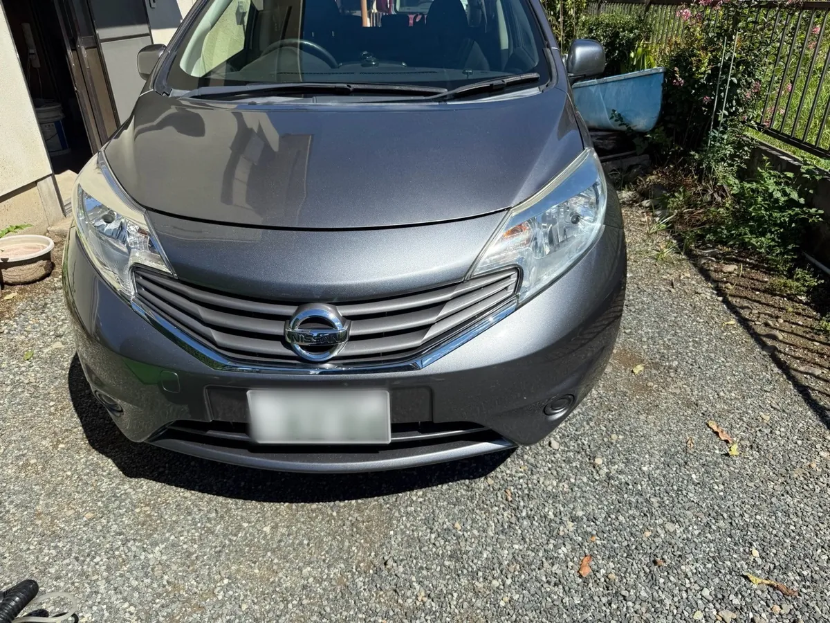 グレーの日産ノートを正面から見たところ。砂利を敷いた駐車場に停まっていて、左手に建物、右手に植え込みと柵がある
