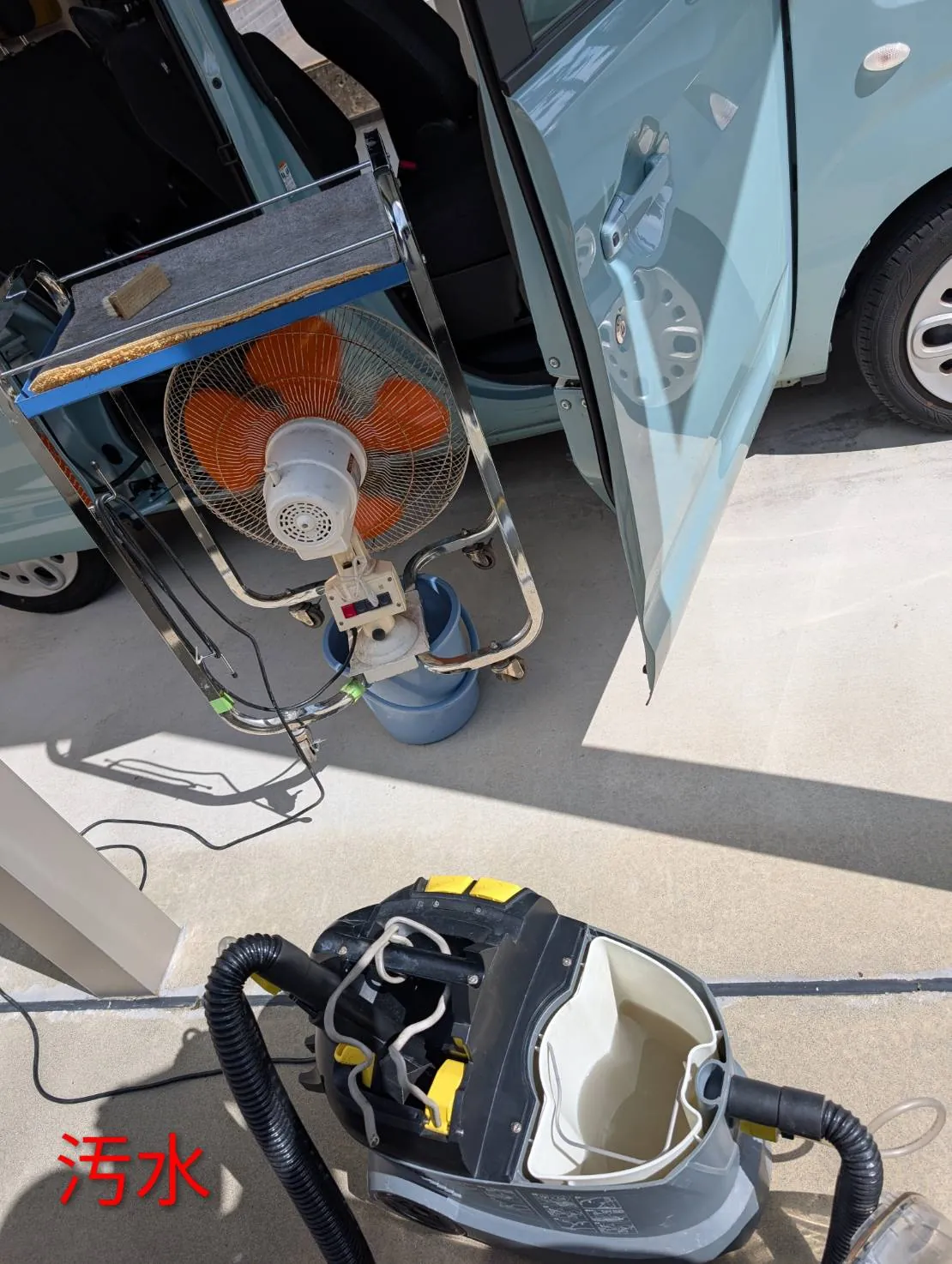 回収した水のタンクを開けたところ。黄色と黒の機械のタンクに濁った水が入っている。奥にステンレスの台車に載せたオレンジ色の羽根の扇風機と青いバケツ、淡い水色のスペーシアの開いたドアとカーポートの床。赤い文字で汚水と入っている