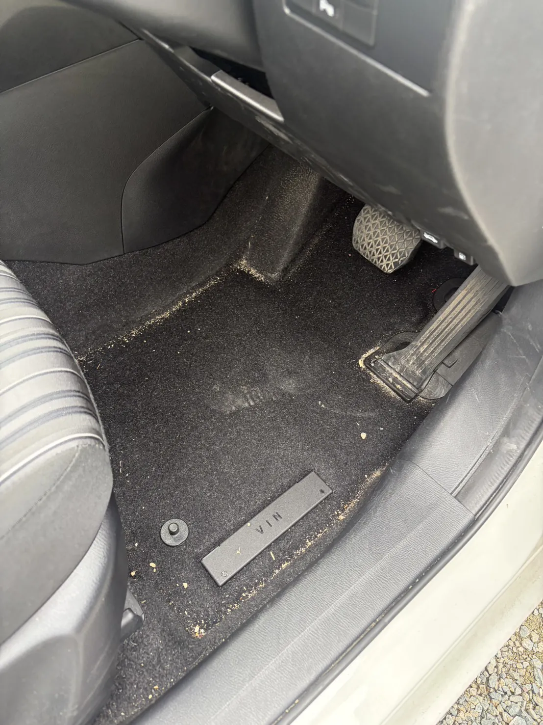 運転席の足元。フロアマットを外した黒いカーペットの上に黄土色の粒が広い範囲に散っている。右奥にアクセルペダルとブレーキペダル、手前にVINと刻印された黒い樹脂のプレートが写っている
