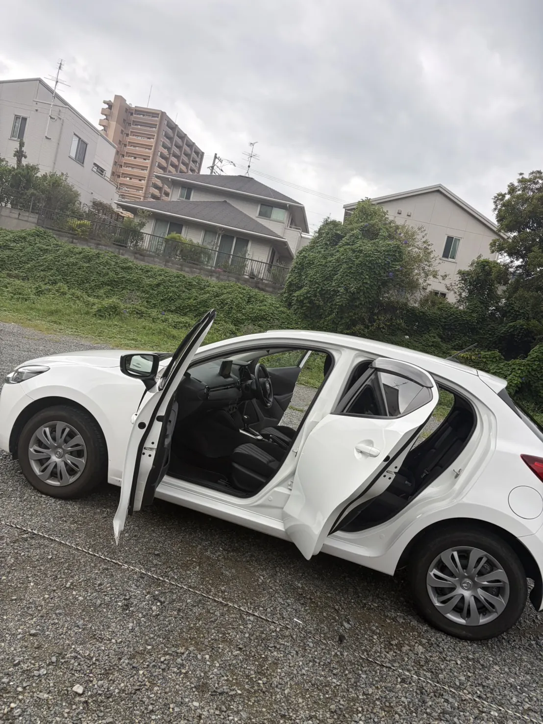 白いマツダのハッチバックを左側から見たところ。運転席のドアと後席のドアを両方開けた状態で、砂利敷きの駐車場に停まっている。背景は住宅と集合住宅