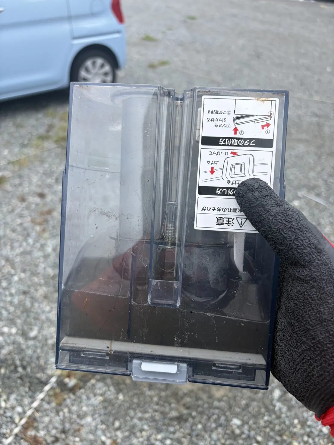 リンサークリーナーの汚水タンクを作業用のグローブをはめた手で持ち上げたところ。透明なタンクの下のほうに黒く濁った水が溜まっている。背景は砂利の地面