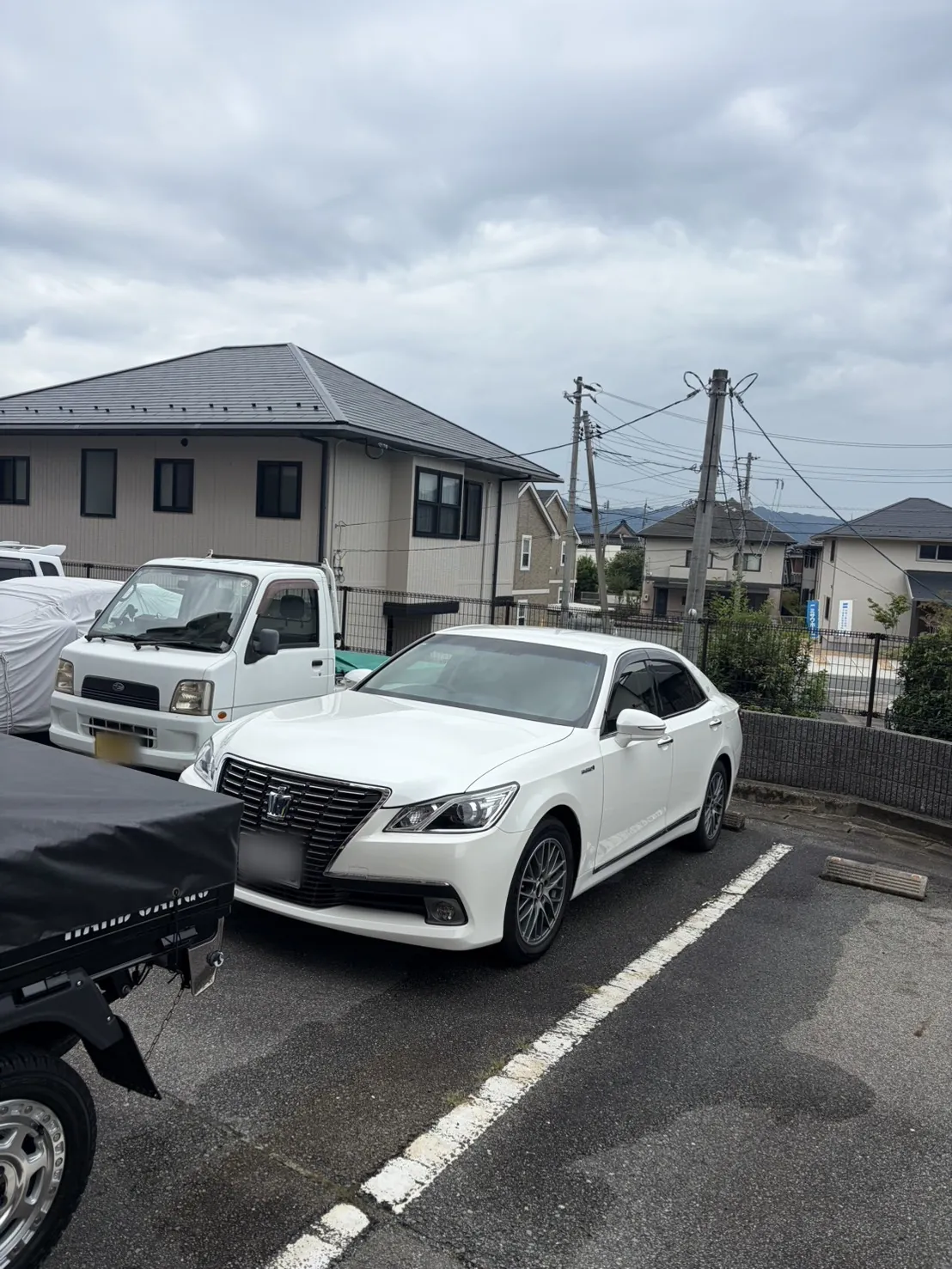 白いトヨタ クラウンを左前から見たところ。舗装された屋外の駐車場に白線に沿って停まっており、左隣に白い軽トラック、背景は住宅街。ナンバープレートはぼかし処理済み