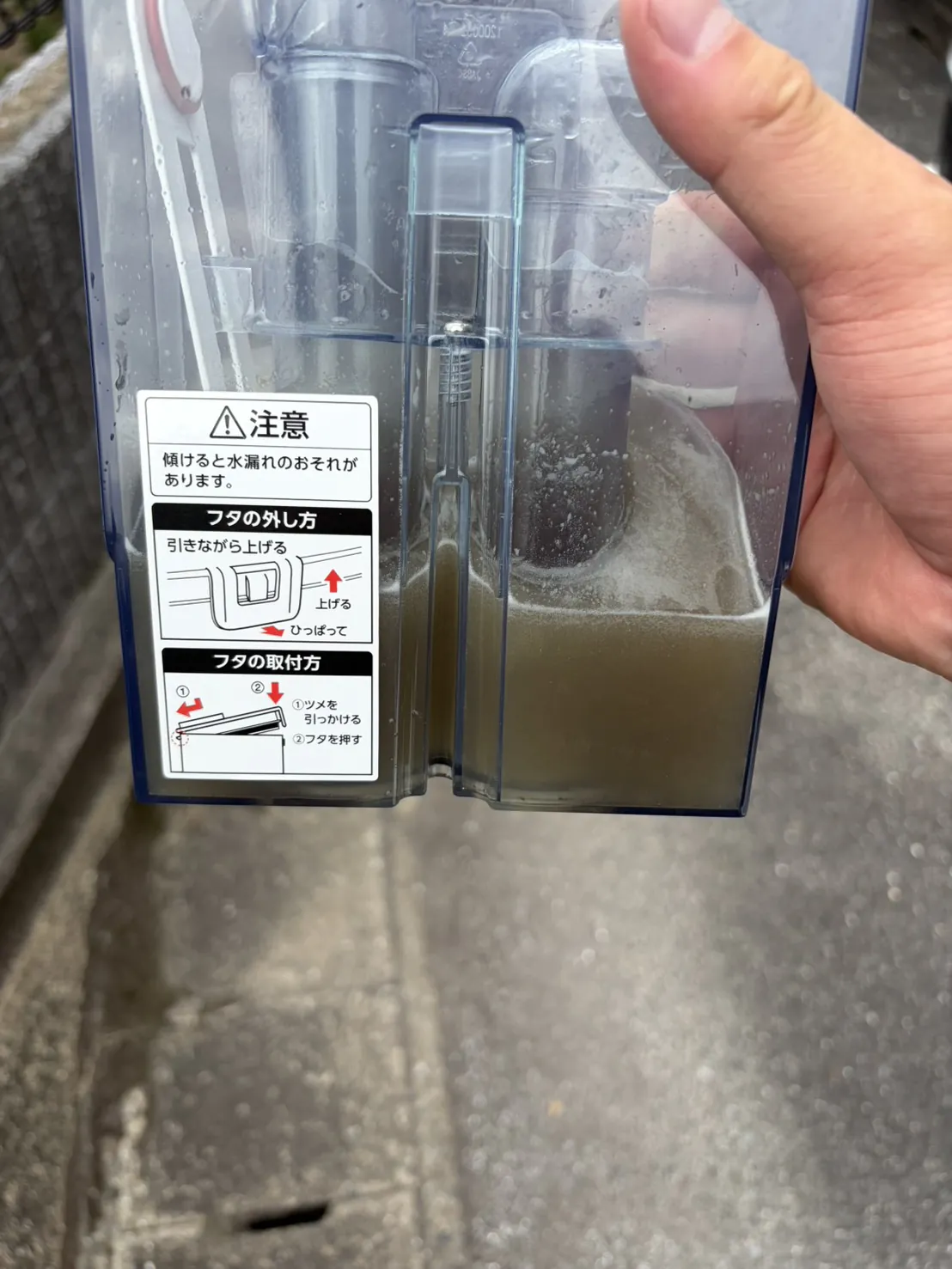 リンサークリーナーの汚水タンクを手で持ち上げたところ。透明なタンクの下半分に茶色く濁った水が溜まり、水面に白い泡の層ができている。背景は屋外のコンクリート