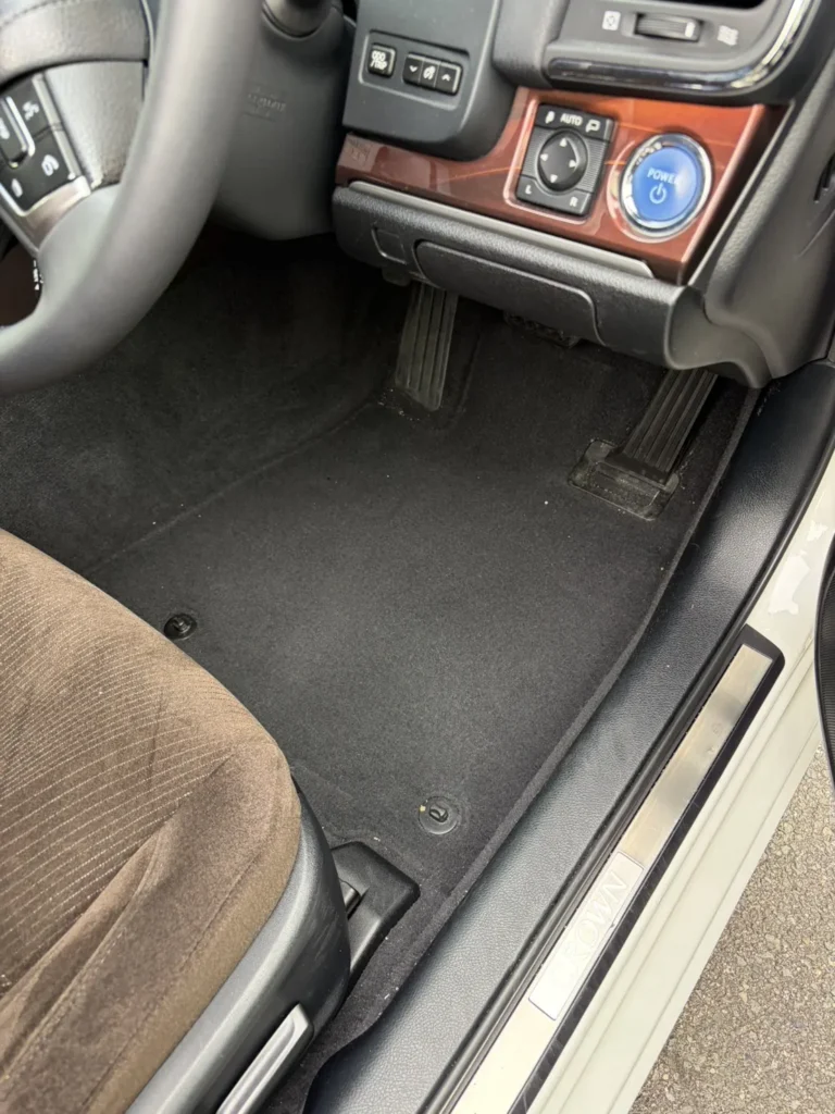 運転席の足元。フロアマットを外した黒いフロアカーペットが出ており、右奥にブレーキペダルとアクセルペダル、右のドアの敷居にCROWNの文字が入った金属のスカッフプレート。中央のパネルは木目調で、丸い青色のPOWERボタンが見える